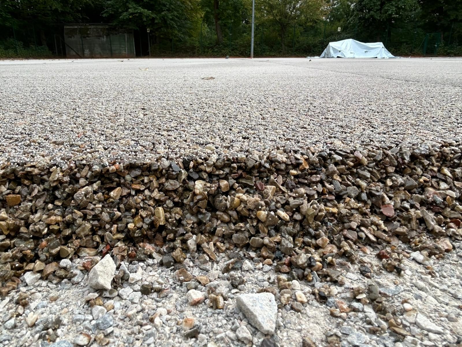 Flacher Blick über einen der hinteren Plätze: vorne die offene Kante aus grobem Kies und Schotter, dahinter die frisch verklebte helle Schicht bis zum Zaun