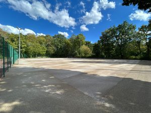 Leerer Tennisplatz mit fertig verlegter Kunststoff-Tragschicht auf einem der hinteren Plätze, im Hintergrund der Baumbestand der Anlage