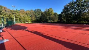 Die hinteren Tennisplätze mit fertig verlegtem rotem HarTru-TopSand-Membranbelag, dahinter Flutlichtmasten, Zaun und Bäume; am Rand stehen Eimer und Werkzeug.