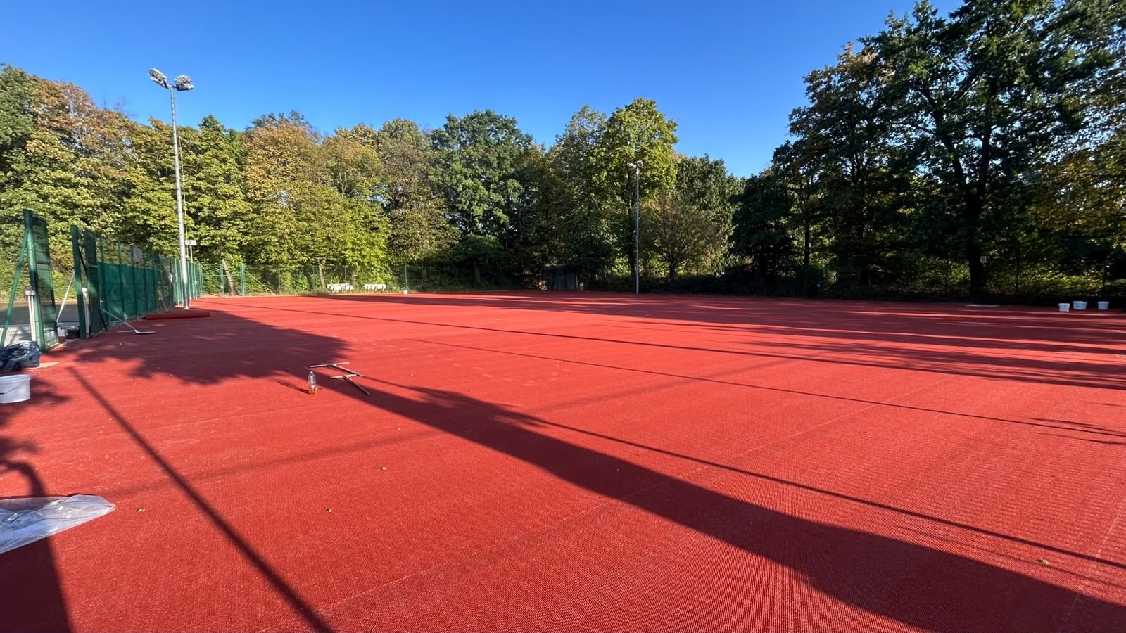 Die hinteren Tennisplätze mit fertig verlegtem rotem HarTru-TopSand-Membranbelag, dahinter Flutlichtmasten, Zaun und Bäume; am Rand stehen Eimer und Werkzeug.