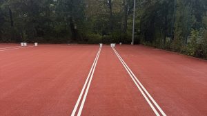 Frisch beschichteter roter Platz P6 mit weiß markierten Linien, am Platzrand stehen Farbeimer, dahinter Zaun und Bäume