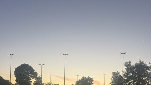 Abenddämmerung über der Tennisanlage mit Flutlichtmasten, Bäumen und laufenden Sprenklern im Hintergrund