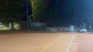 Tennisplatz nachts mit Flutlicht, im Hintergrund gestapelte Sandsäcke am Zaun