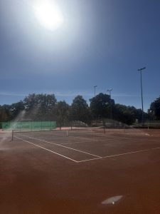 Einer der neuen Plätze mit Netz und frisch markierten Linien in der Sonne, die Bewässerungssprenkler laufen.
