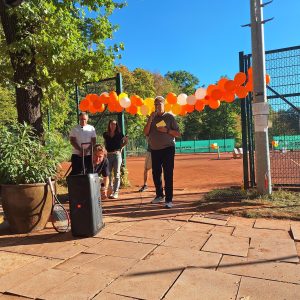 Ansprache bei der Eröffnungsfeier: Ein Mann spricht am Eingang zu den Plätzen ins Mikrofon, über ihm eine Girlande aus orangefarbenen und weißen Luftballons, im Hintergrund ein neuer Platz.
